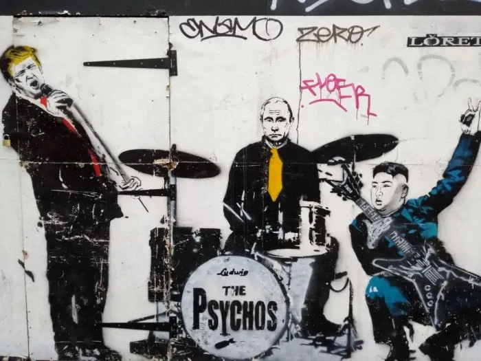 Art work on a wall. Probably stencil and spray paint. A band is playing. 3 members: singer, guitarist, drummer. They pose like a punk band. From the kick-drum print, we can tell the band is called "The Psychos". Each member is wearing a suit or a costume and look like Donald Trump on the mic, Valdimir Putin on the drum and Kim Jong Un on the lead guitar.