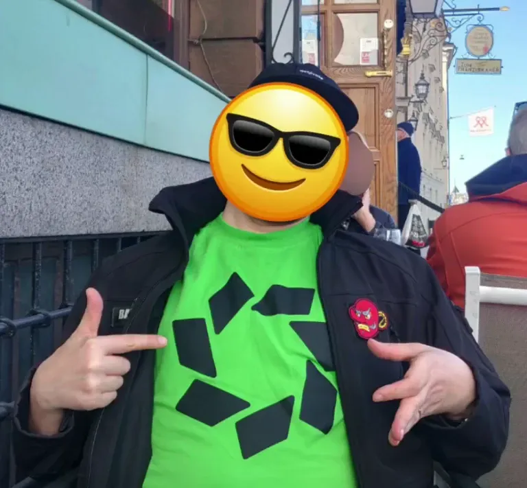 A dyne who's face is hidden behind an emoji, pointing to their bright green t-shirt with a logo looking like a stylized bunghole