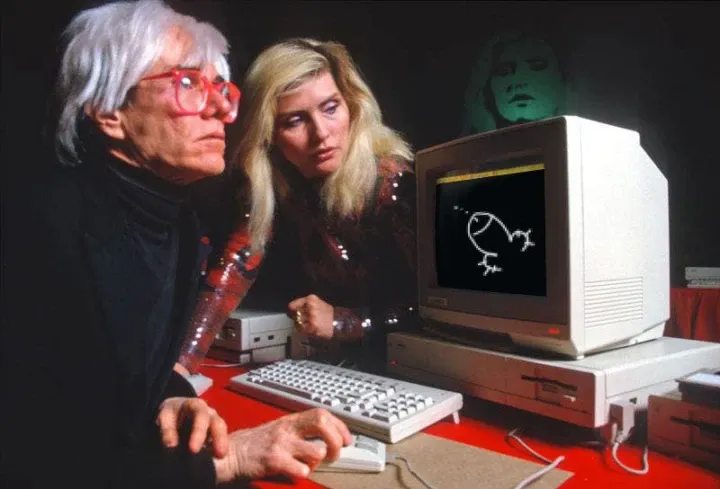 Andy Warhol and Blondie in front of an old Macintosh computer. Warhol holds the mouse and blondie is peeking at the screen. On the screen a classic depiction of a dick and balls.